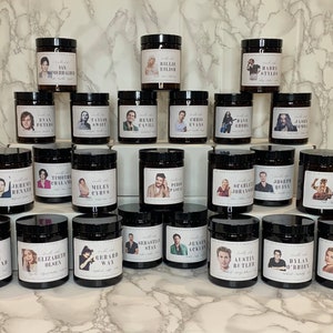 May include: A collection of 30 brown glass candle jars with black lids. Each jar features a different celebrity's face on the label. The candles are arranged in three rows of ten.