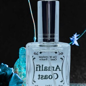 Puede incluir: Una botella de vidrio transparente de perfume Coast Amalfi con una tapa plateada. La botella est&aacute; etiquetada con "Coast Amalfi" y "The Marvelous Candle Studio" en una fuente de escritura negra. La botella est&aacute; sobre una superficie azul brillante.
