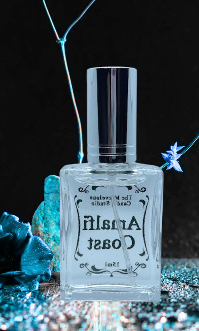 Puede incluir: Una botella de vidrio transparente de perfume Coast Amalfi con una tapa plateada. La botella est&aacute; etiquetada con "Coast Amalfi" y "The Marvelous Candle Studio" en una fuente de escritura negra. La botella est&aacute; sobre una superficie azul brillante.
