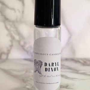 May include: A clear glass roll-on bottle with a black cap and a white label. The label has a black and white illustration of angel wings and the text "Daryl Dixon" with the words "Gift of Hand-Crafted Scents" below.