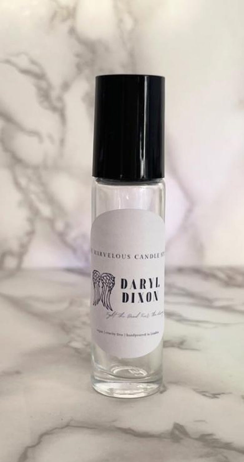 May include: A clear glass roll-on bottle with a black cap and a white label. The label has a black and white illustration of angel wings and the text "Daryl Dixon" with the words "Gift of Hand-Crafted Scents" below.