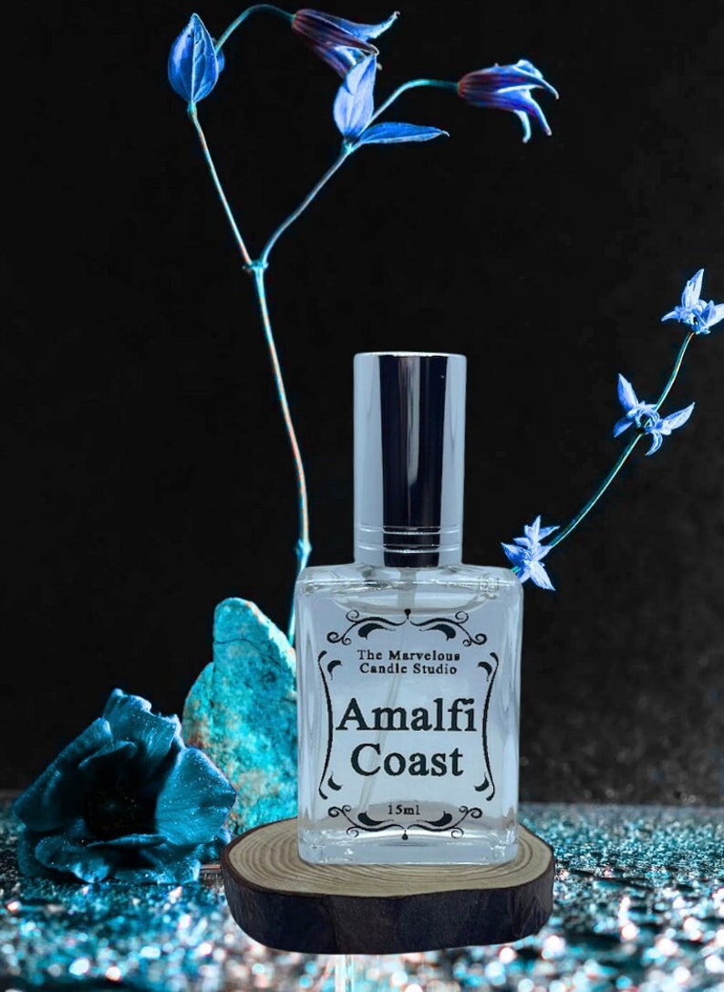 Puede incluir: Una botella de vidrio transparente con una tapa plateada y una etiqueta que dice "The Marvelous Candle Studio Amalfi Coast 15ml". La botella est&aacute; sentada sobre una rodaja redonda de madera. El fondo es negro con flores azules.
