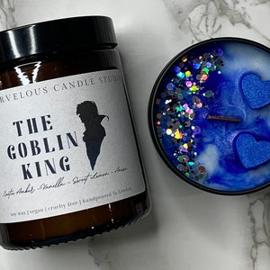 Könnte beinhalten: Ein braunes Glasgefäß mit einer Kerze und einem weißen Etikett, auf dem "The Marvelous Candle Studio" und "The Goblin King" mit der Silhouette eines Mannes mit Hut steht. Die Kerze ist aus Sojawachs, vegan, tierversuchsfrei und handgefertigt in London. Die Kerze befindet sich neben einer blauen und weißen Kerze mit Glitzer und zwei blauen herzförmigen Dekorationen.