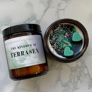 May include: Two brown glass jar candles. One is closed with a black lid and a label that reads "THE KINGDOM OF TERRASEN" with scent details. The other is open, revealing a white wax base with green accents, glitter, and heart-shaped wax melts.