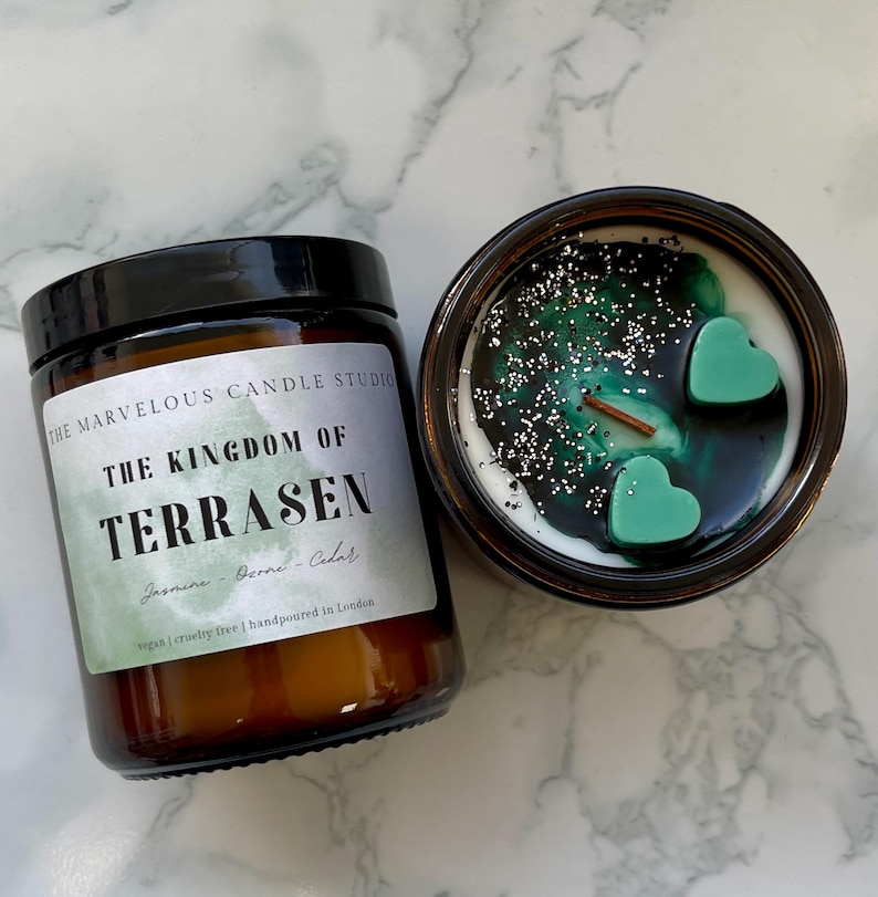 May include: Two brown glass jar candles. One is closed with a black lid and a label that reads "THE KINGDOM OF TERRASEN" with scent details. The other is open, revealing a white wax base with green accents, glitter, and heart-shaped wax melts.