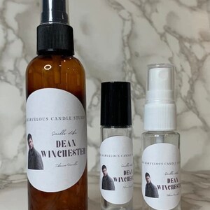 Op de afbeelding: Drie glazen flessen met etiketten waarop "Smells Like Dean Winchester" staat en een foto van een man te zien is. De flessen zijn van verschillende grootte en hebben zwarte en witte doppen.
