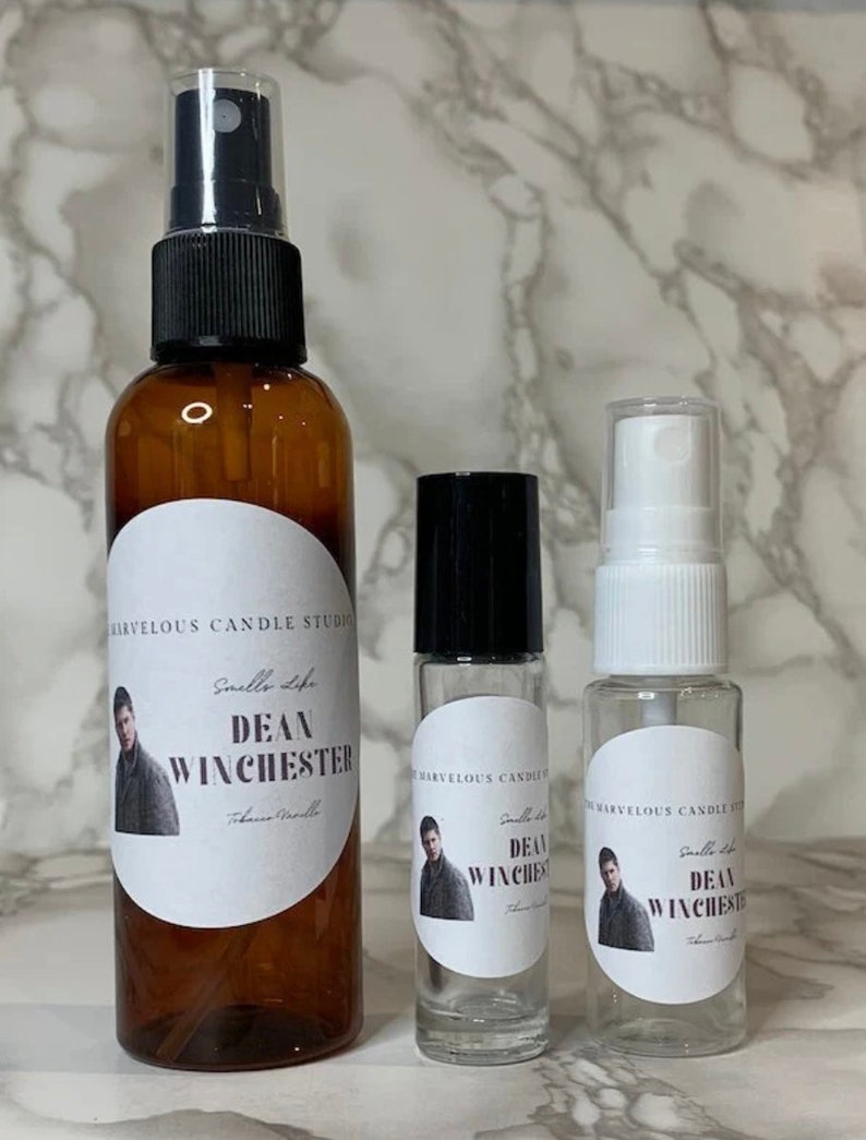 Op de afbeelding: Drie glazen flessen met etiketten waarop "Smells Like Dean Winchester" staat en een foto van een man te zien is. De flessen zijn van verschillende grootte en hebben zwarte en witte doppen.