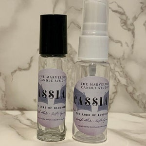 May include: Two clear glass bottles with black and white labels. The labels have a purple background and say "The Marvelous Candle Studio" and "Cassia - The Lord of Bloodroot Bath & Body - Cassia Spice" in a black, stylized font. The bottles are on a white marble surface.