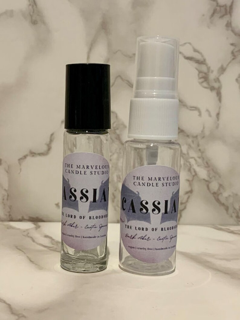 May include: Two clear glass bottles with black and white labels. The labels have a purple background and say "The Marvelous Candle Studio" and "Cassia - The Lord of Bloodroot Bath & Body - Cassia Spice" in a black, stylized font. The bottles are on a white marble surface.