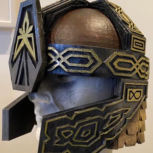 Peut inclure: Un casque noir et or avec des motifs géométriques complexes. Le casque a un sommet arrondi et un masque facial avec un motif géométrique. Le casque est fait d'un matériau dur et a une surface texturée.