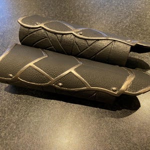 May include: Two black leather arm guards with silver-colored trim and decorative stitching. The arm guards are designed with a textured surface and are likely for costume or cosplay purposes.