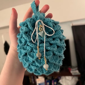 May include: A teal blue crocheted drawstring bag with a white cord and a gold charm. The bag has a beaded tassel with light blue beads and gold spacers.