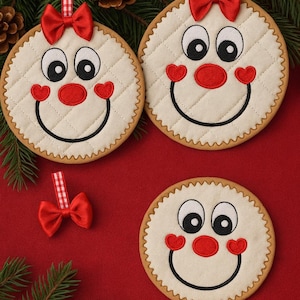 Può includere: Tre decorazioni natalizie rotonde color crema con facce sorridenti, nasi rossi e guance a forma di cuore. Ognuna ha un fiocco rosso in cima e un bordo smerlato marrone. Le decorazioni sono su uno sfondo rosso con aghi di pino e pigne.