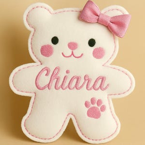 Può includere: Un giocattolo di peluche bianco a forma di orsetto con dettagli ricamati in rosa. L'orso ha occhi neri, guance rosa e un fiocco rosa. Il nome "Chiara" è ricamato in rosa, insieme a un'impronta di zampa.