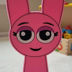 May include: A pink felt cutout shaped like a cartoon character with long rabbit-like ears. The character features large black eyes with white pupils, pink eyelids, black eyelashes, and a simple black curved line for a mouth, all outlined in black.