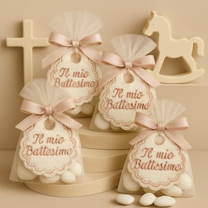 Può includere: Quattro piccoli sacchetti trasparenti pieni di caramelle bianche, ciascuno legato con un nastro rosa e un'etichetta con la scritta "Il mio Battesimo". Una croce bianca e un cavallo a dondolo sono sullo sfondo. I sacchetti sono su un espositore a più livelli.