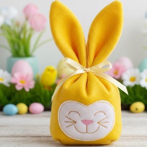 Può includere: Un sacchetto regalo giallo a forma di coniglietto pasquale con un viso bianco, naso rosa e baffi ricamati. La borsa ha orecchie alte e dritte ed è legata con un nastro color crema. Lo sfondo include decorazioni pasquali.