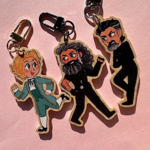 May include: Three wooden keychains featuring cartoon characters. The first keychain features a character with blonde hair wearing a blue and green outfit. The second keychain features a character with a beard and black hair wearing a black outfit. The third keychain features a character with black hair wearing a black outfit.