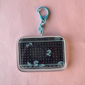 Może przedstawiać: Przezroczysty akrylowy brelok z turkusowym zapięciem i łańcuszkiem. Prostokątna zawieszka ma czarne tło z turkusowymi cyframi i symbolami. U góry widnieje napis "LUMON". Brelok znajduje się na różowym tle.