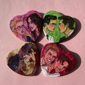 Può includere: Quattro spille a forma di cuore con illustrazioni di coppie. Le spille presentano opere d'arte colorate con un effetto olografico. Le coppie sono raffigurate in varie pose, con diversi colori della pelle e dei capelli. Lo sfondo è rosa.