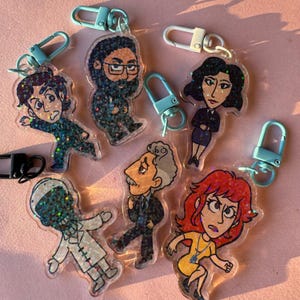 May include: Six colorful glitter keychain charms featuring cartoon characters. Each charm has a blue and white metal clasp. The characters include a man with a beard, a woman with dark hair, a man with a mustache, a woman with red hair, a man with a black and white face, and a man with brown hair.