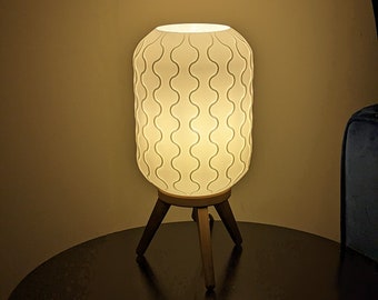 Op de afbeelding: Een witte tafellamp met een patroonkap en een houten driepoot. De lamp straalt een warm licht uit, waardoor een gezellige sfeer ontstaat. De kap heeft een herhalend golvend ontwerp. De lamp staat op een donkere ronde tafel.
