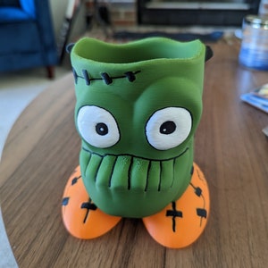 May include: Green Frankenstein monster-shaped ceramic pencil holder with orange feet.