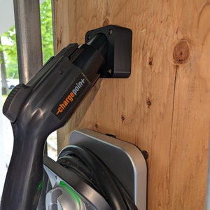 Puede incluir: Una estación de carga para vehículos eléctricos con un conector negro y una base plateada. El conector, etiquetado como "chargepoint+", está montado en una superficie de madera.