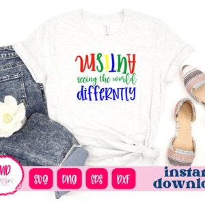 May include: A white t-shirt with the text "Autism seeing the world differently" in rainbow colors. The shirt is on a light gray surface with blue jeans and a white flower.