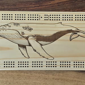Peut inclure: Un jeu de cribbage en bois avec un dessin gravé d'une baleine. La planche rectangulaire est bordée de trous percés pour les chevilles. Le mot "Skunk" est imprimé sur le côté gauche.