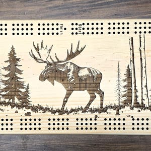 Peut inclure: Jeu de cribbage en bois avec un motif gravé au laser d'un orignal dans une forêt. La planche rectangulaire a des coins arrondis et est marquée de nombreux trous pour les chevilles. Les mots "Start" et "Finish" sont gravés.