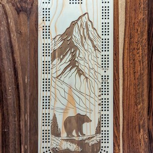 Peut inclure: Plateau de cribbage en bois avec un motif de montagne et d'ours. Le plateau est de couleur brun clair et présente une scène de montagne en noir et blanc avec un ours brun au premier plan. Le plateau est doté d'une grille de trous pour les pions et les mots "Départ" et "Arrivée" sont imprimés sur le côté.