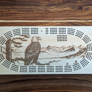 Peut inclure: Un plateau de jeu de cribbage en bois avec un motif gravé au laser d'un aigle chauve perché sur une branche surplombant un lac de montagne. Le plateau a une finition en bois naturel et des points noirs pour le score.