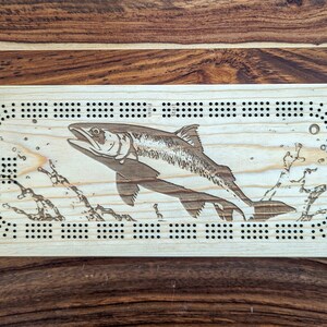 Peut inclure: Un plateau de cribbage en bois avec un motif de saumon. Le plateau a une finition en bois naturel et présente une gravure détaillée d'un saumon sautant hors de l'eau. Le plateau a une disposition traditionnelle de cribbage avec des chevilles et des trous de pointage.