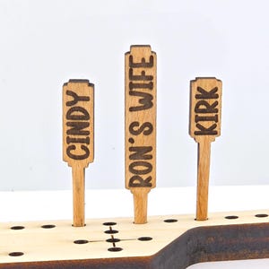 May include: A wooden game board with three wooden pegs. The pegs have the names "Cindy", "Ron's Wife", and "Kirk" written on them.