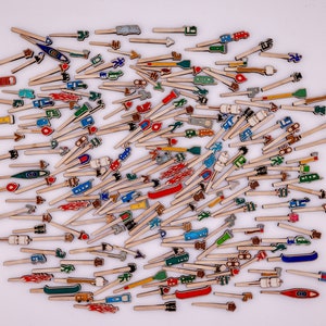 May include: A collection of small, wooden arrow-shaped objects with colourful painted designs. The designs include cars, boats, arrows, and other symbols. The objects are scattered on a white surface.