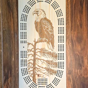 Peut inclure: Un plateau de jeu de cribbage en bois avec un motif découpé au laser d'un aigle chauve perché sur une branche de pin. Le plateau comporte une rangée circulaire de trous pour marquer les points.