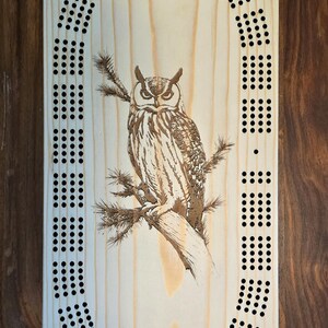 May include: A wooden cribbage board with a brown owl design in the center. The board has a circular track of holes around the owl, and a smaller square of holes above the owl.