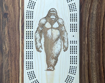 May include: A wooden cribbage board featuring a laser-etched design of a gorilla. The board has a circular track with holes for pegs.