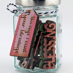 May include: A glass jar with a silver lid filled with wooden blocks that spell out "blessings". A pink and white tag attached to the jar reads "Always count your blessings".