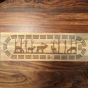 Peut inclure: Plateau de cribbage en bois sur le thème de la faune. Le plateau présente une piste circulaire avec des trous pour les pions et une scène de cerfs et d'élans dans un décor de forêt.
