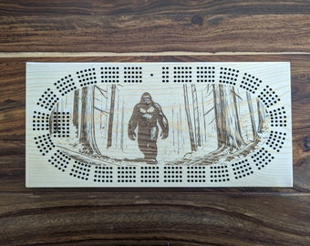 May include: A wooden cribbage board featuring a laser-engraved image of Bigfoot walking through a forest. The board has a circular playing area with holes for pegs.