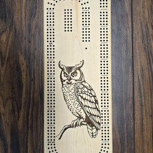 May include: A wooden cribbage board with a detailed engraving of an owl perched on a branch. The board features a classic track layout with numerous holes for peg placement, and is set against a dark wood background.