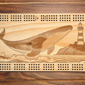 May include: A wooden cribbage board with a whale and lighthouse design. The board has a light wood finish and features a whale jumping out of the water near a lighthouse on a rocky island. The board has a grid of holes for pegs and the word "Stunk" is printed on the board.