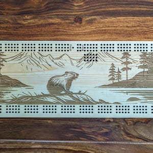 May include: A wooden cribbage board with a mountain and lake scene featuring a beaver. The board has a light wood finish and black dots for scoring. The text "Finish" and "Shunk" are visible on the board.