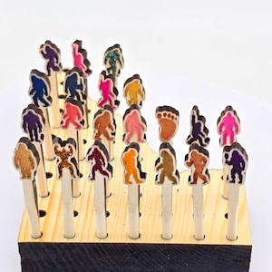 May include: A wooden stand with 24 holes holds colorful, glittery, wooden Bigfoot figures. The figures are in various poses and colors, including pink, purple, green, orange, and brown.