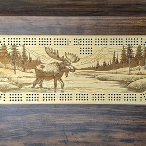 Peut inclure: Jeu de cribbage en bois avec une scène de wapiti gravée. Le jeu présente un paysage détaillé avec un wapiti dans une rivière, entouré d'arbres et de montagnes. Il comporte des trous pour les chevilles et est en bois clair.