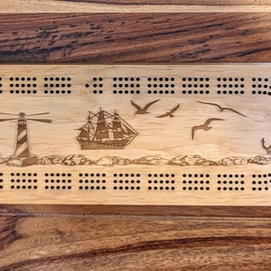 May include: A wooden cribbage board with a nautical theme. The board features a lighthouse, a ship, seagulls, and an anchor. The board has a light brown finish and black dots for scoring.