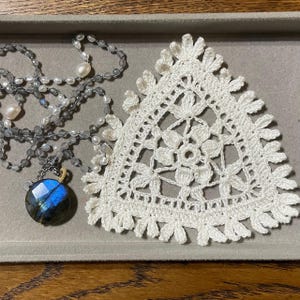 May include: A necklace with a blue and green gemstone pendant and a beaded chain, alongside a white crocheted doily with a floral design. The necklace features pearls and gray beads. The doily is triangular with a scalloped edge.