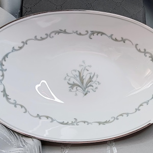 May include: Oval white serving dish with a silver rim and a decorative gray floral and scroll design. The center features a stylized floral arrangement. A heart-shaped design is on the left side of the dish.
